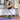 A woman in a black sports bra and light gray leggings performs a squat on a purple yoga mat, wearing a green ankle support band with a black logo patch. The setting is a bright, modern kitchen with white cabinets and wooden floors.