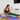 A woman in a black sports bra and light blue leggings sits on a purple yoga mat, stretching her leg while using a green fabric stretch band with a small black XPEED label.