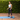 A woman in a light blue sports bra and black leggings stands on a blue yoga mat outdoors, holding a pink resistance band in front of a green hedge.