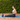A woman in a light blue sports bra and black leggings sits on a blue Xpeed exercise mat outdoors, using a pink Xpeed resistance tube with a black handle against a green hedge background.