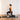 A woman in a black strapless top and black leggings holds a gray xpeed kettlebell while performing a lunge on a dark yoga mat in a room with white curtains and a light wood floor.