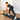 A woman in a black sports bra and leggings does a bent over row with an xpeed soft kettlebell on a black yoga mat in a bright room with white curtains and wood floors.